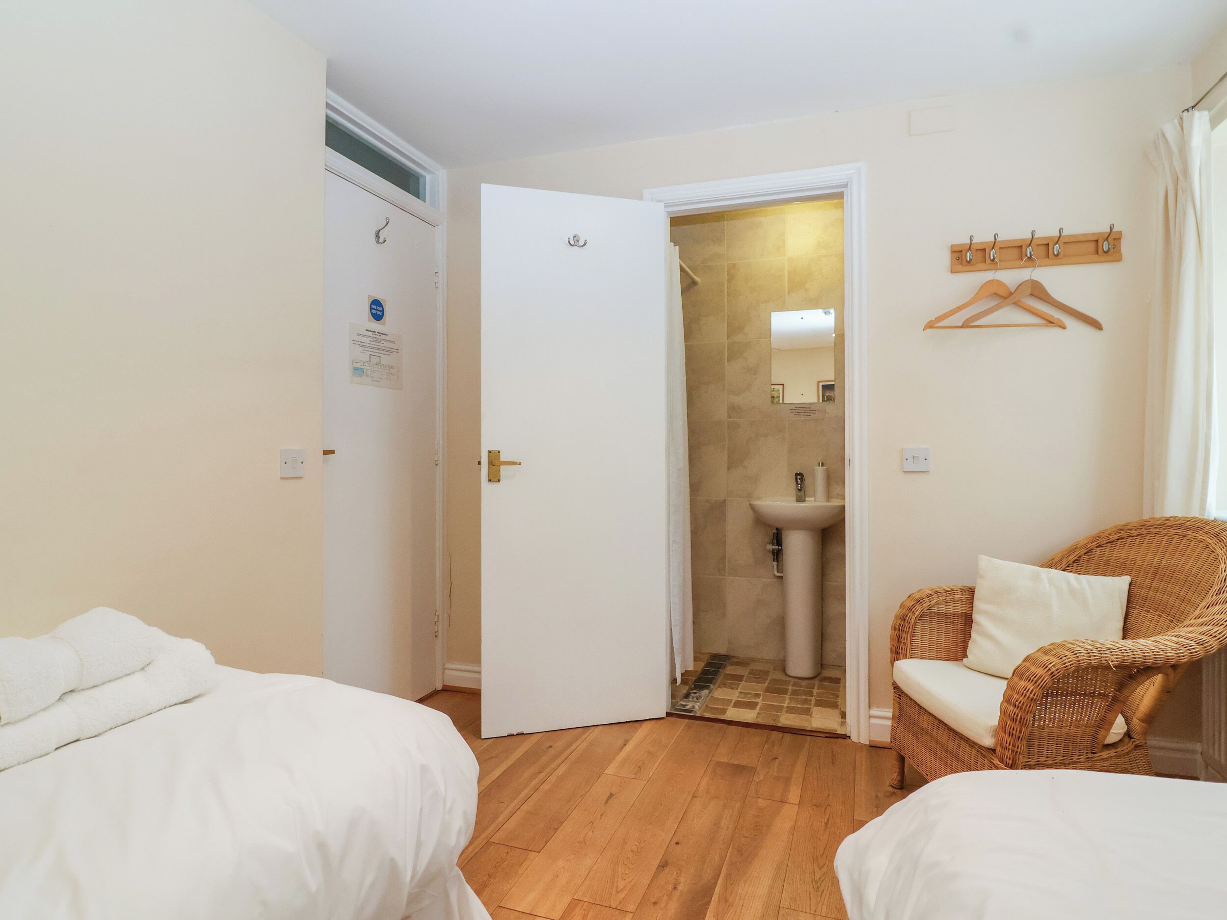 A hotel room with two beds, a wicker chair, a bathroom with a sink and mirror, and a coat rack.