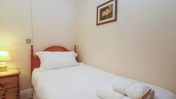 Een klein, net bed met witte lakens en een houten hoofdbord, een nachtkastje met een lamp, en een ingelijst foto aan de muur.