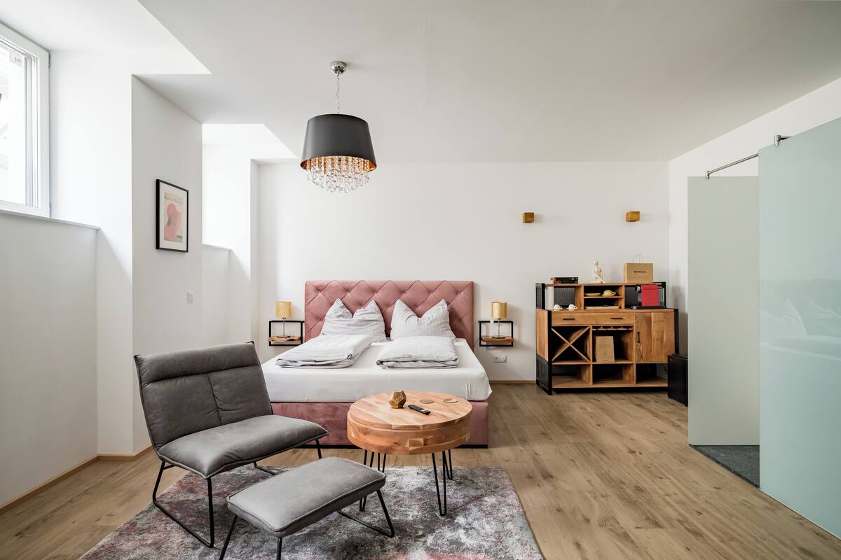A modern bedroom with a large bed, a grey armchair, a small round table, and a hanging light fixture.