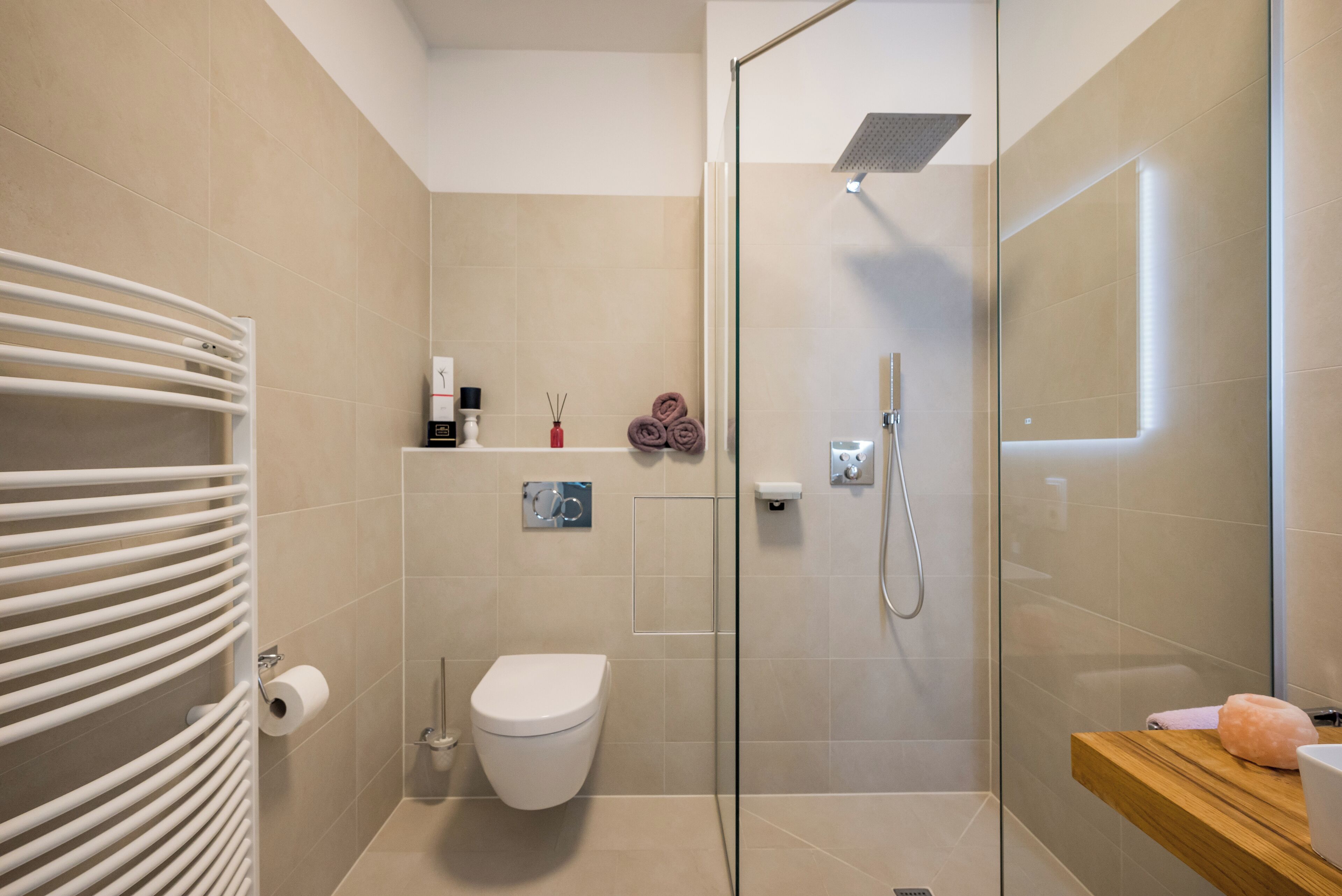 Ein modernes Badezimmer mit Duschkabine aus Glas, Toilette, Handtuchhalter und einer Holzablage mit einem rosafarbenen Gegenstand.