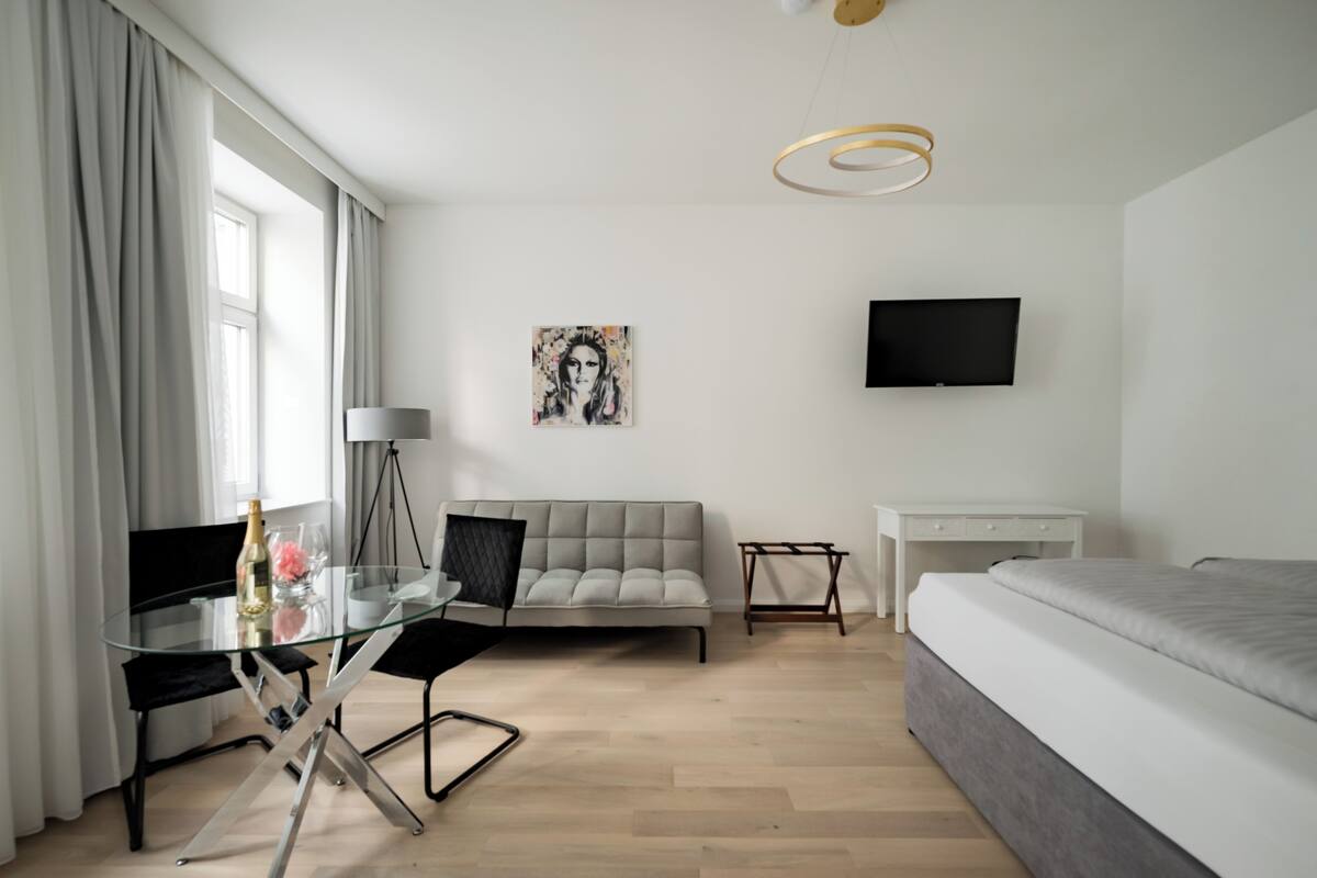A modern hotel room with a glass dining table, a sofa, a bed, a TV, and a painting on the wall.