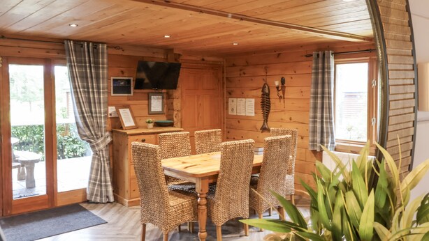 En stuga byggd av trä med ett matbord och stolar, en tv som är monterad på väggen och ett fönster med gardiner.