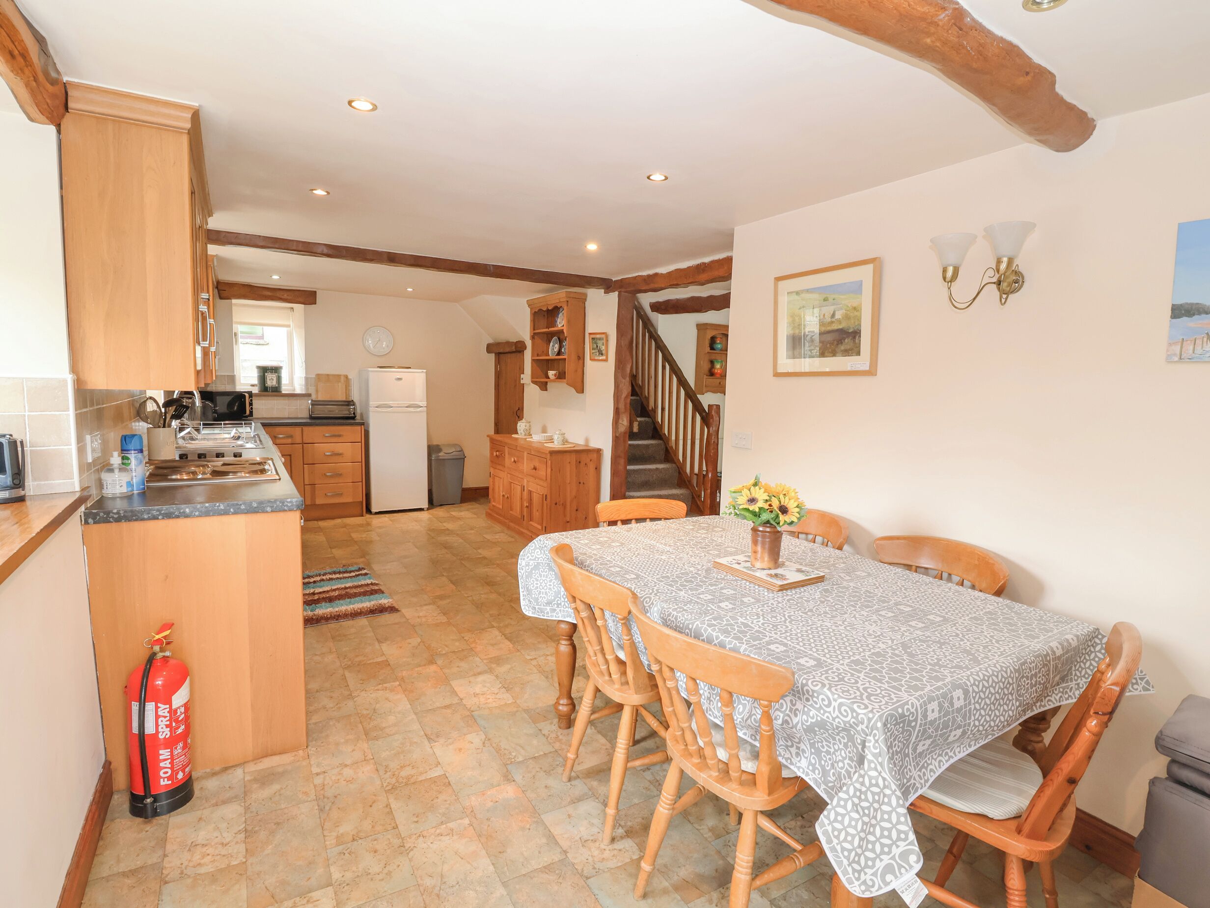 A cozy kitchen with wooden cabinets, a dining table with chairs, a fire extinguisher, and a staircase leading to another room.