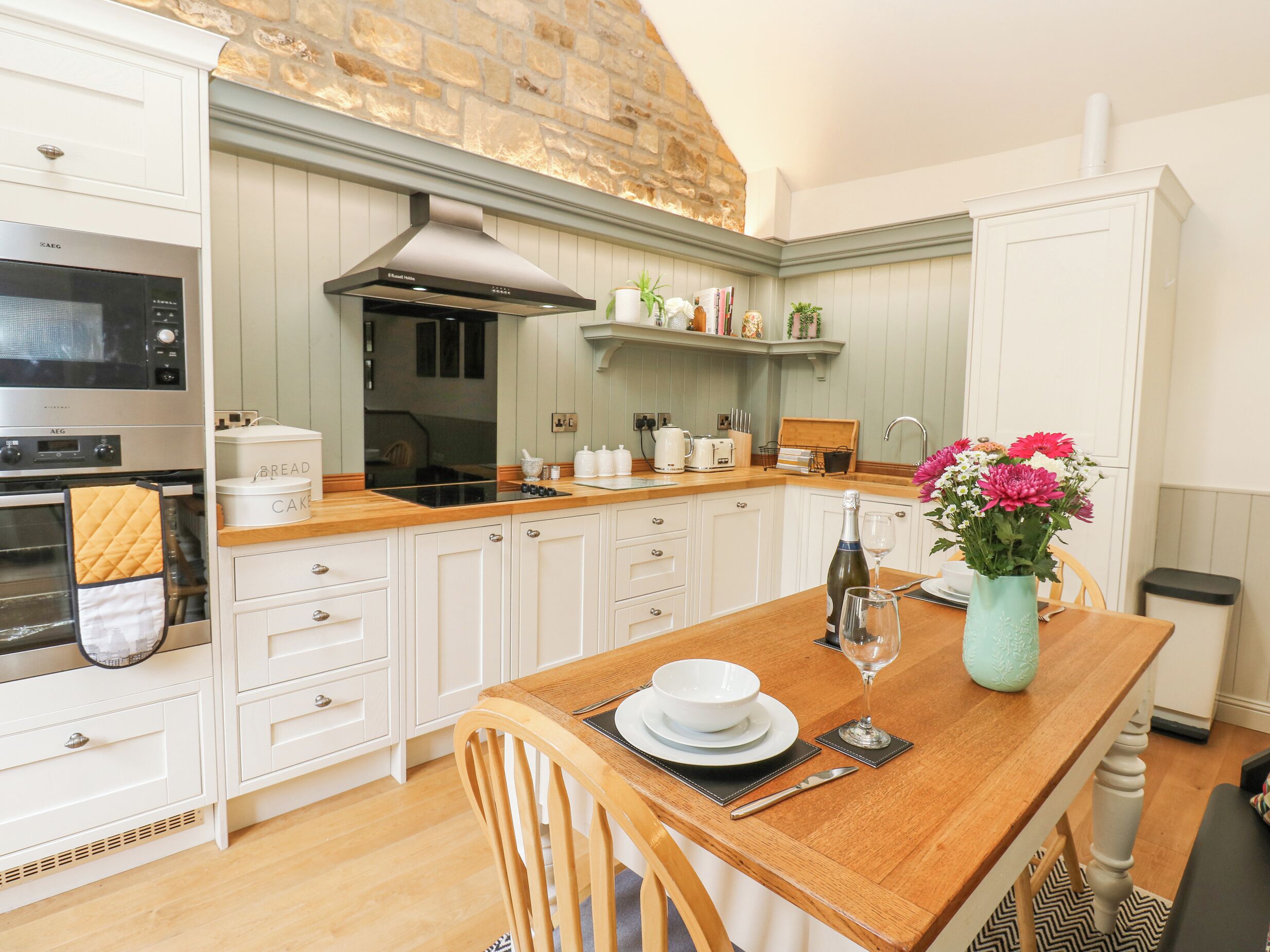 A kitchen with white cabinets, a wooden countertop, a built-in oven, a stove, and a dining table set with plates, a wine glass, and a vase with flowers.