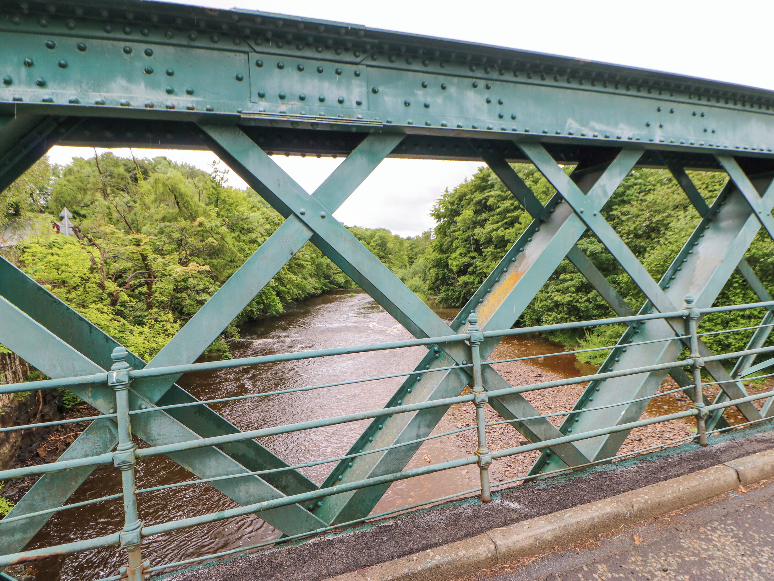 A green metal bridge with a river underneath and trees in the background.