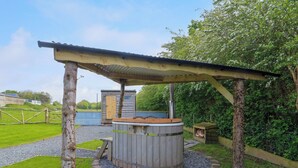 A rustic outdoor hot tub area with a wooden structure overhead, surrounded by greenery and a gravel path. - Llety Llanfair (Caernarfon)