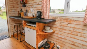 A compact kitchen with wooden walls, a small refrigerator, a coffee maker, and a kettle on the counter. - Llety Llanfair (Caernarfon)