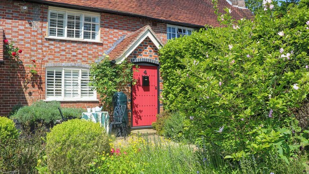 Eine rote Tür zu einem Haus aus Ziegelsteinen, umgeben von Grünpflanzen und einem Garten.
