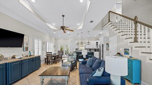 A spacious living room with a large sofa, a wooden coffee table, and a dining area with chairs. There is a kitchen in the background with white cabinets and a staircase leading to another level.