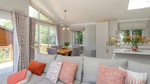A modern living room with a grey sofa, orange and patterned cushions, a dining area with a table and chairs, and a kitchen with white cabinets. - Waterside Lodge (Windermere)