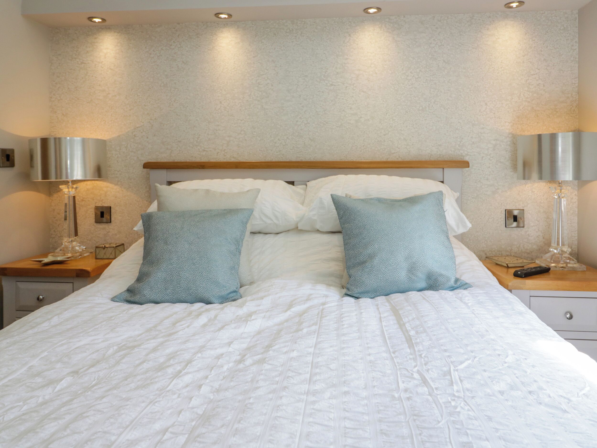 A neatly made bed with white bedding and two matching pillows, flanked by two bedside tables with lamps and a remote control.
