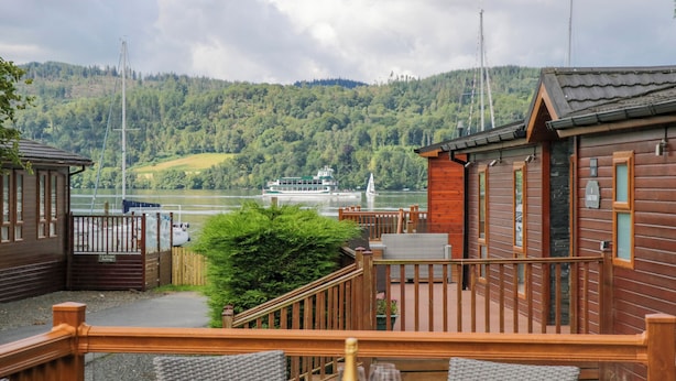 A wooden cabin with a deck overlooking a lake, a boat in the distance, and a forested hillside. - Waterside Lodge (Windermere)