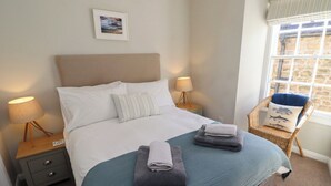 A neatly made bed with a blue bedspread, two pillows, and folded towels. There are two bedside tables with lamps, a wicker chair with a cushion, and a framed picture on the wall. - Aln Cottage (Alnwick)