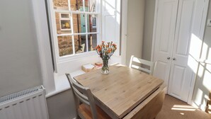 A dining area with a wooden table, two chairs, a vase with flowers, and a window with a view of a brick building. - Aln Cottage (Alnwick)