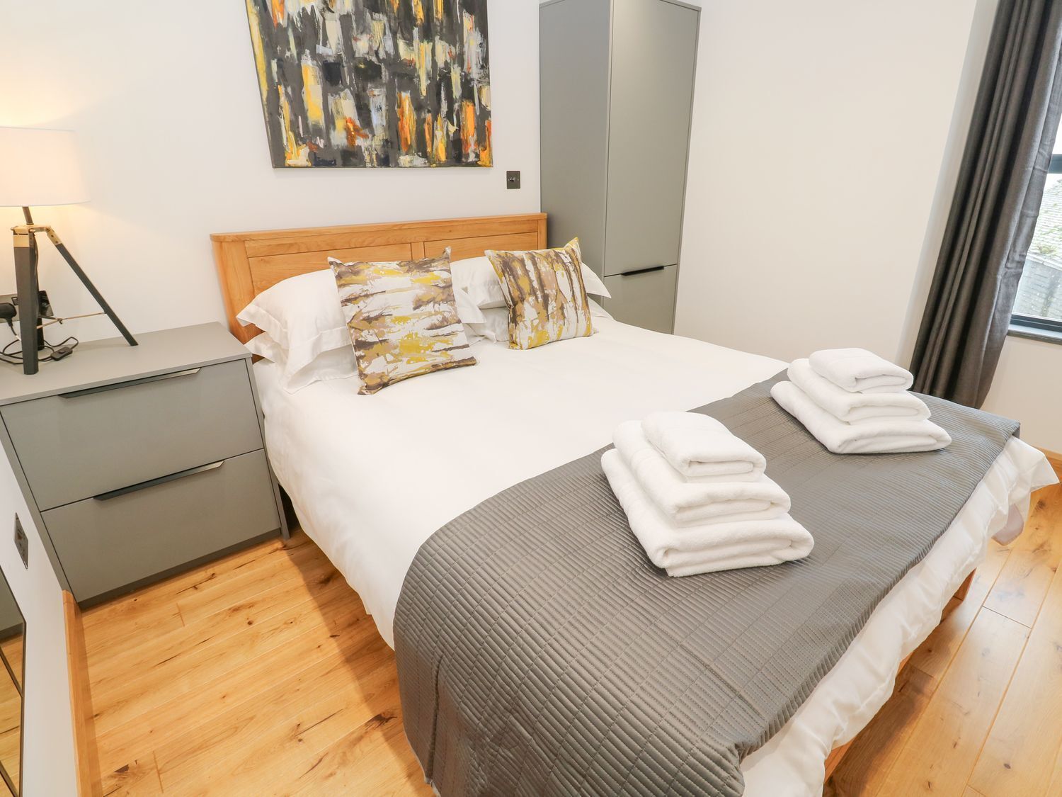 Una habitación de hotel moderna con piso de madera, cama con cubrecama a rayas gris y blanco, varios almohadones, mesita de noche con lámpara y un cuadro en la pared.