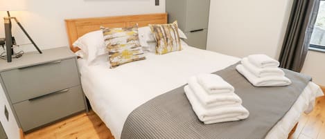 Una habitación de hotel moderna con piso de madera, cama con cubrecama a rayas gris y blanco, varios almohadones, mesita de noche con lámpara y un cuadro en la pared.