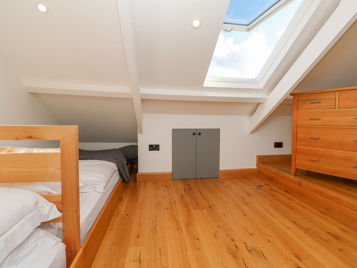 Un dormitorio con tragaluz, piso de madera, cama, armario y un mueble gris.