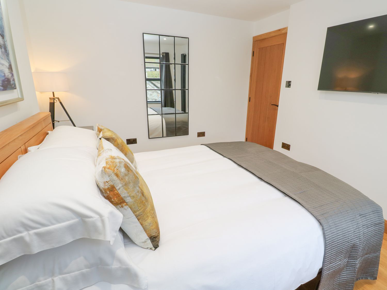 A hotel room with a bed, a bedside lamp, a mirror, a door, and a TV.
