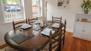 A dining room with a wooden table set for six, surrounded by chairs. There are windows with blinds, a wall-mounted light fixture, and a framed picture above the table. - 8 Nicholas Court (Chester)
