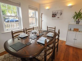 A dining room with a wooden table set for six, surrounded by chairs. There are windows with blinds, a wall-mounted light fixture, and a framed picture above the table.