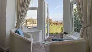 A room with two white armchairs, a window with curtains, and a view of a grassy area and a stone wall. - Blue Yonder (Weymouth)