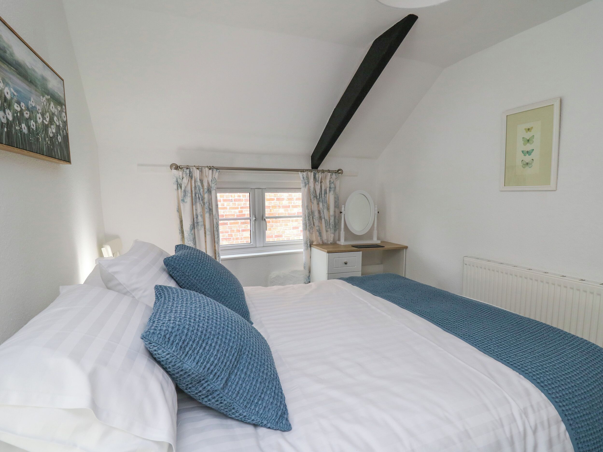 A bedroom with a bed, a window with curtains, a bedside table with a mirror, and a radiator.