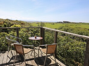 A wooden deck with two wicker chairs and a small round table, overlooking a green landscape with fields and distant hills. - Ty Mynydd (Holyhead)
