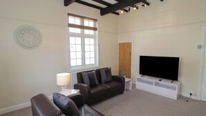 A living room with a brown leather sofa, a white lamp, a flat-screen TV on a white stand, and a window with blinds. - Above The Pub (Birmingham)