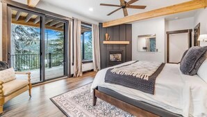 Een slaapkamer met een groot bed, een plafondventilator, een openhaard en een balkon met uitzicht op bomen.