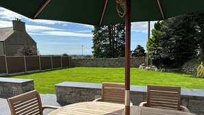 Un patio exterior con una mesa redonda de madera y cuatro sillas de madera, rodeado por un muro de piedra y un césped verde.
