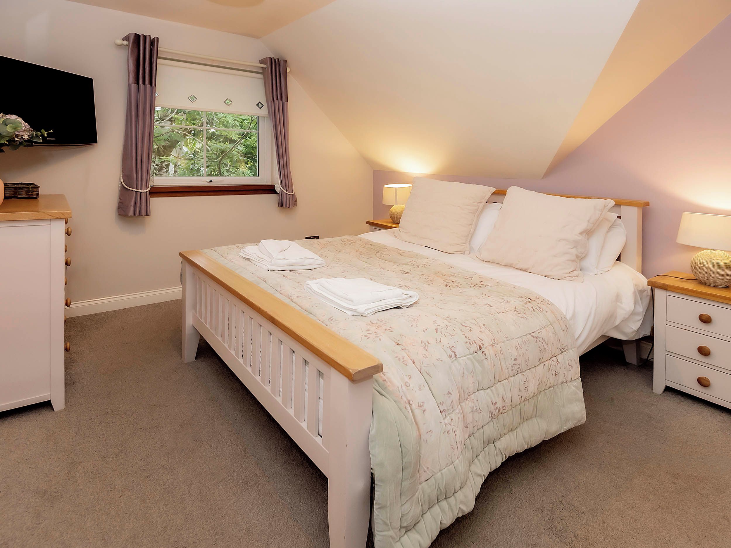 A bedroom with a bed, bedside lamps, a window with curtains, a TV, and a bedside table.