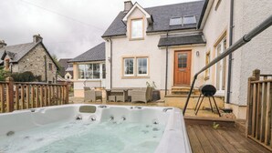Un bain à remous dans un jardin, avec une terrasse en bois, un barbecue et une maison à la façade en pierre et à la clôture en bois.