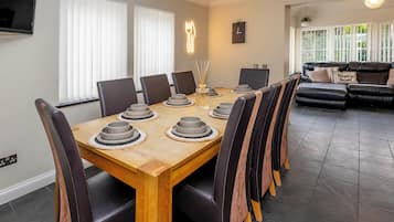 Uma sala de jantar com mesa de madeira posta para seis pessoas, rodeada por cadeiras de couro, uma TV na parede e um relógio.