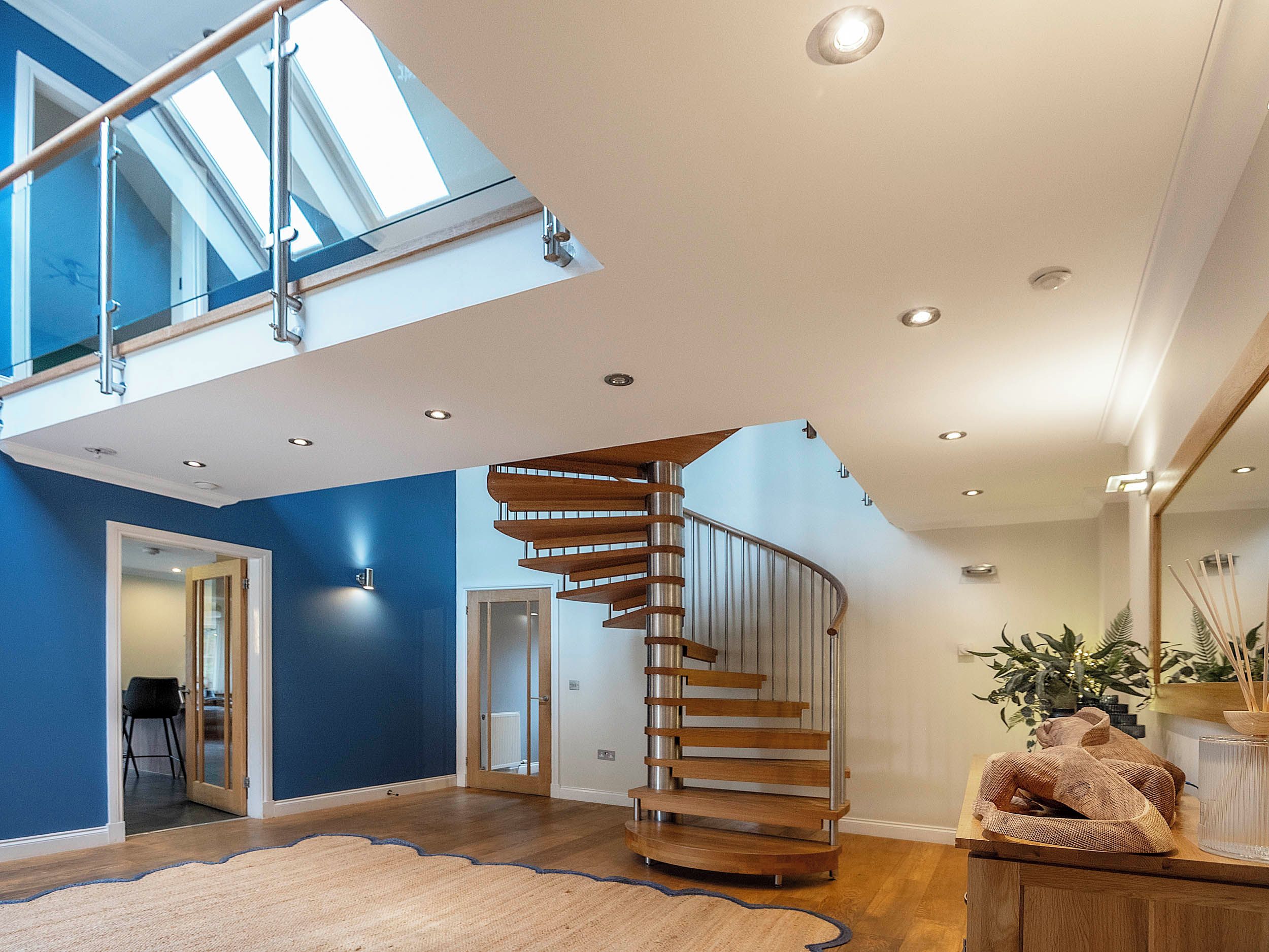 Een modern interieur met een houten spiltrap, een hoog plafond met dakramen en een grote spiegel die de ruimte weerspiegelt.