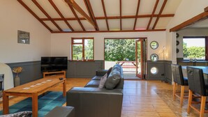 A spacious living area with a sofa, a TV, and a wooden table. There are large windows and a view of greenery outside. - Bay Tree Lodge 35 (Ilfracombe)