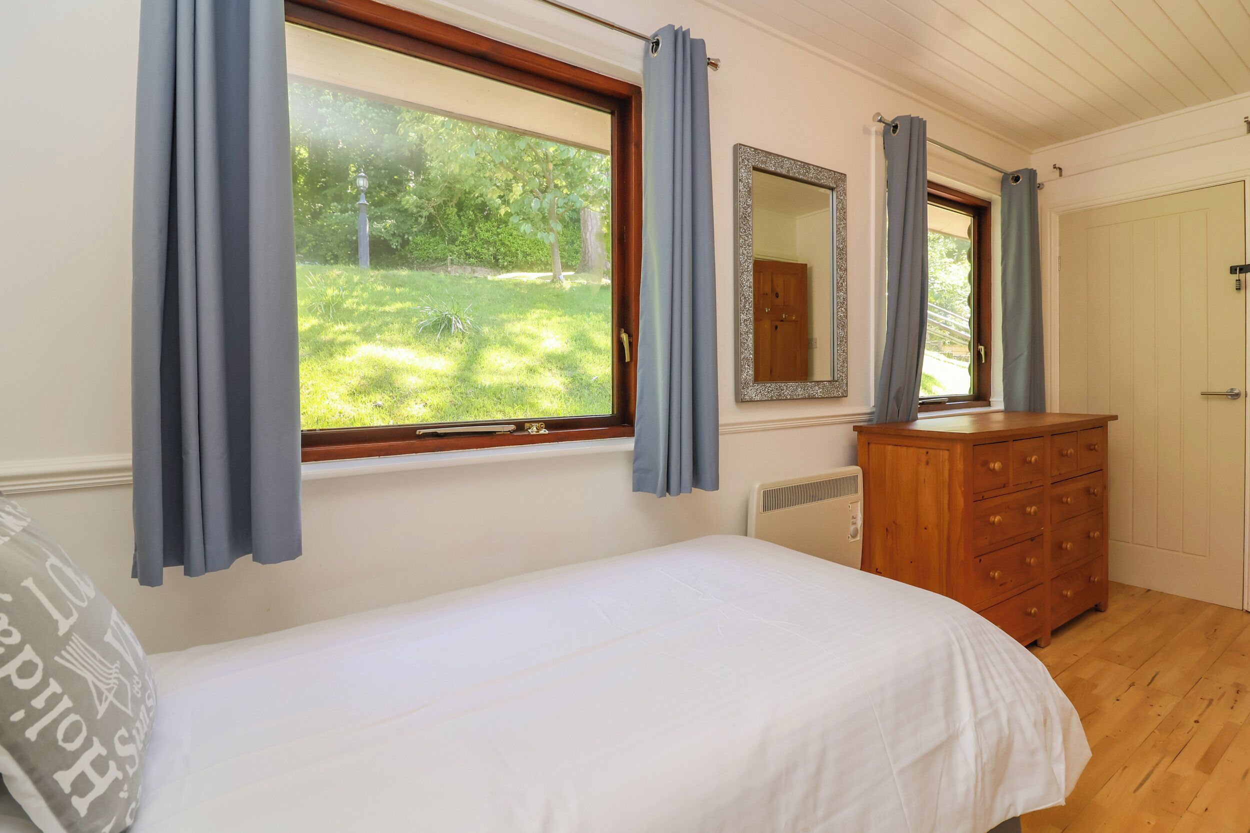 A bedroom with a single bed, a dresser, a mirror, and a window with curtains.