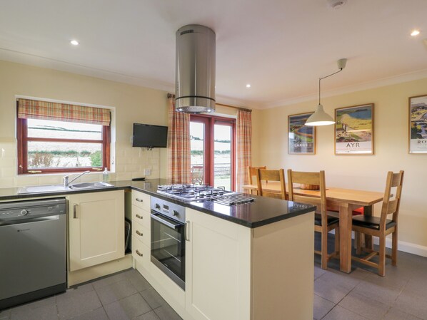 Une cuisine compacte équipée d’une cuisinière, d’un four et d’un lave-vaisselle, un coin repas avec des chaises en bois et une grande fenêtre avec des rideaux.