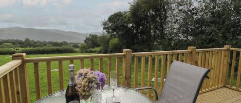 A round glass table with a bottle, a glass, and a vase of flowers on a wooden deck with a view of a green field and trees.