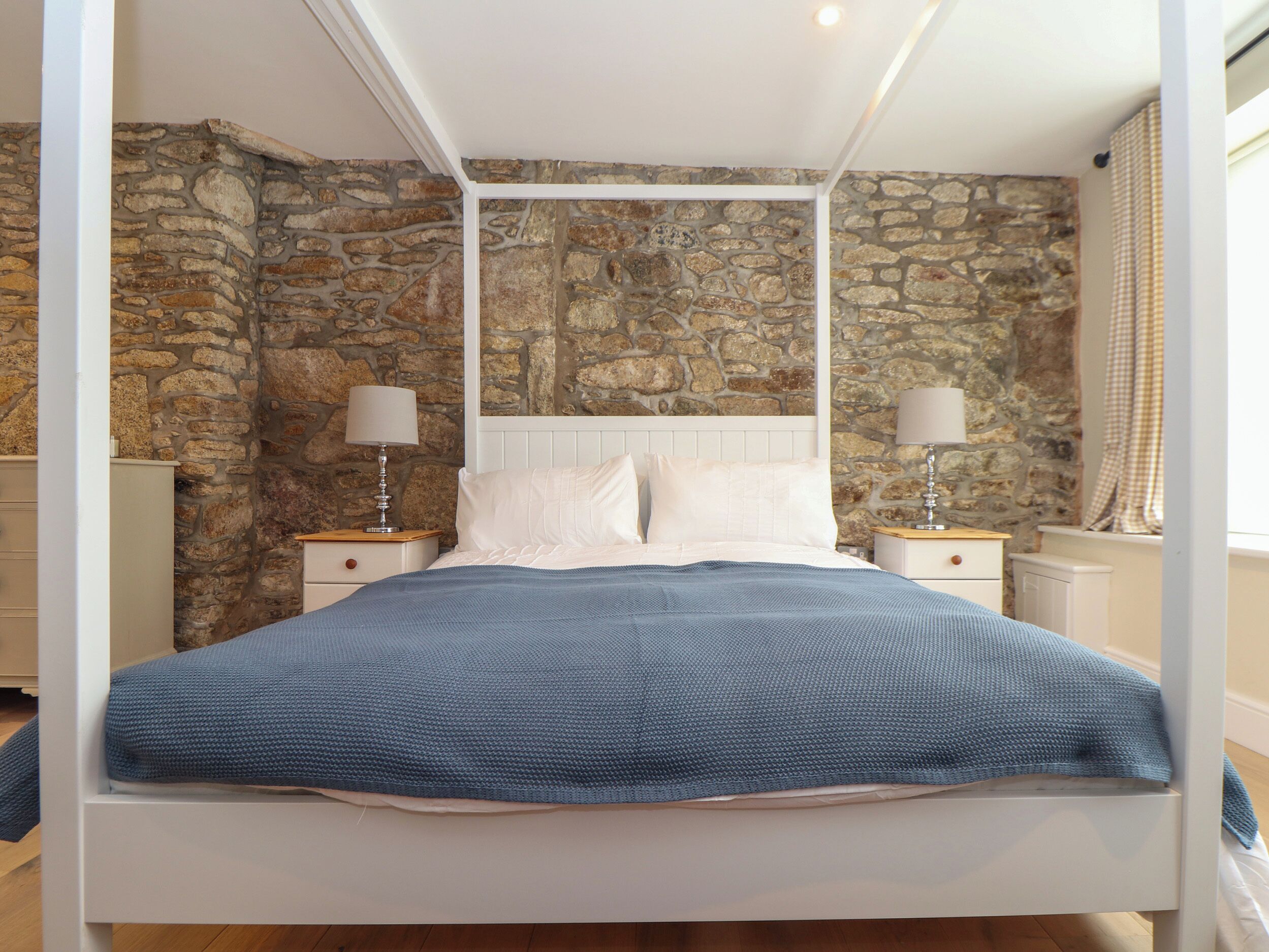 A four-poster bed with white bedding and a blue throw, set against a stone wall backdrop.