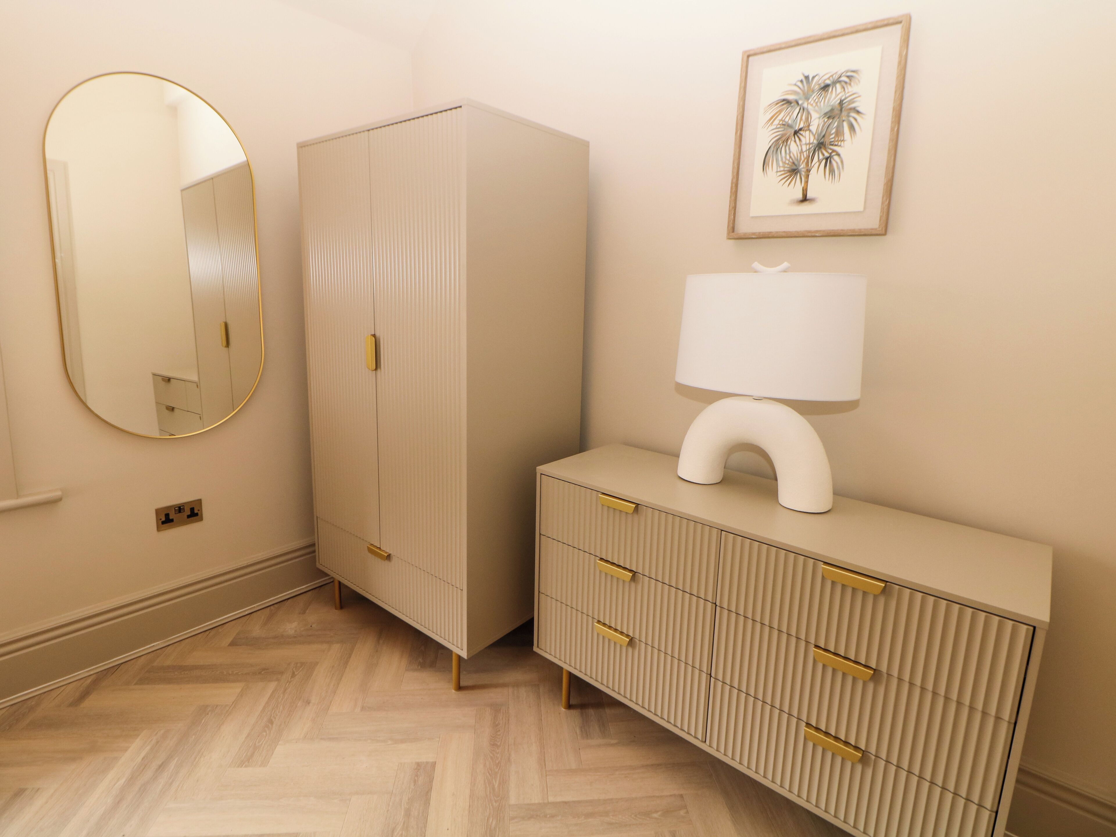 Una habitación con armario, espejo, mesita de noche con lámpara y un cuadro en la pared.
