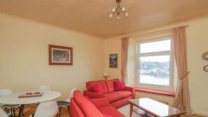 A living room with a red sofa, a glass coffee table, and a dining table with chairs. There is a window with a view of a town and a clock on the wall. - Flat 2 Beechcroft (Dartmouth)