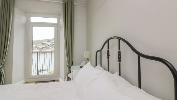 A bedroom with a bed, a nightstand, a lamp, and a view of a balcony with curtains. - Flat 2 Beechcroft (Dartmouth)