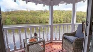 Un espacio al aire libre techado con terraza de madera, mobiliario de mimbre y vistas a un río y un bosque.