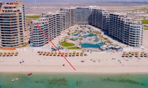 A large hotel with multiple high-rise buildings, a central pool area, and a beach with sun loungers.