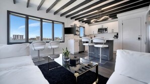 Un apartamento moderno con sala de estar, cocina y comedor. Hay ventanas amplias, una alfombra negra y muebles blancos.