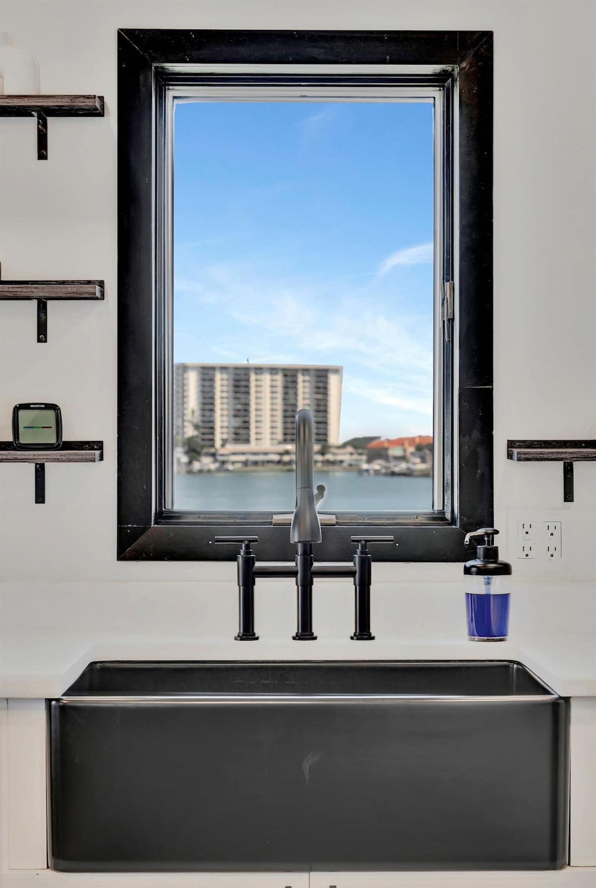 A kitchen with a black sink, a window with a view of buildings and water, and a small clock on the wall.