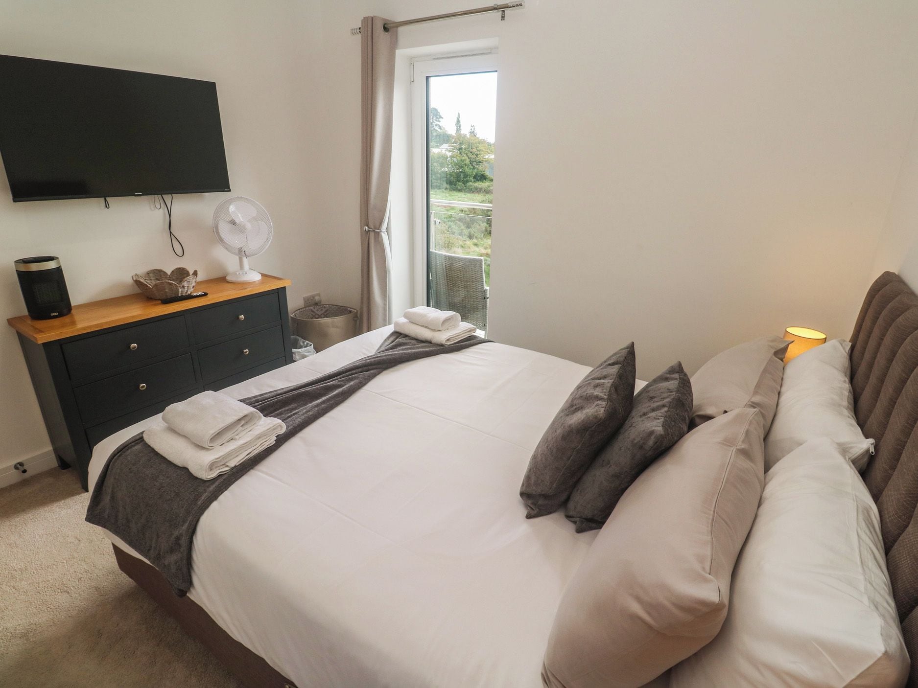 A bedroom with a bed, bedside table, lamp, and a view of greenery through a window.