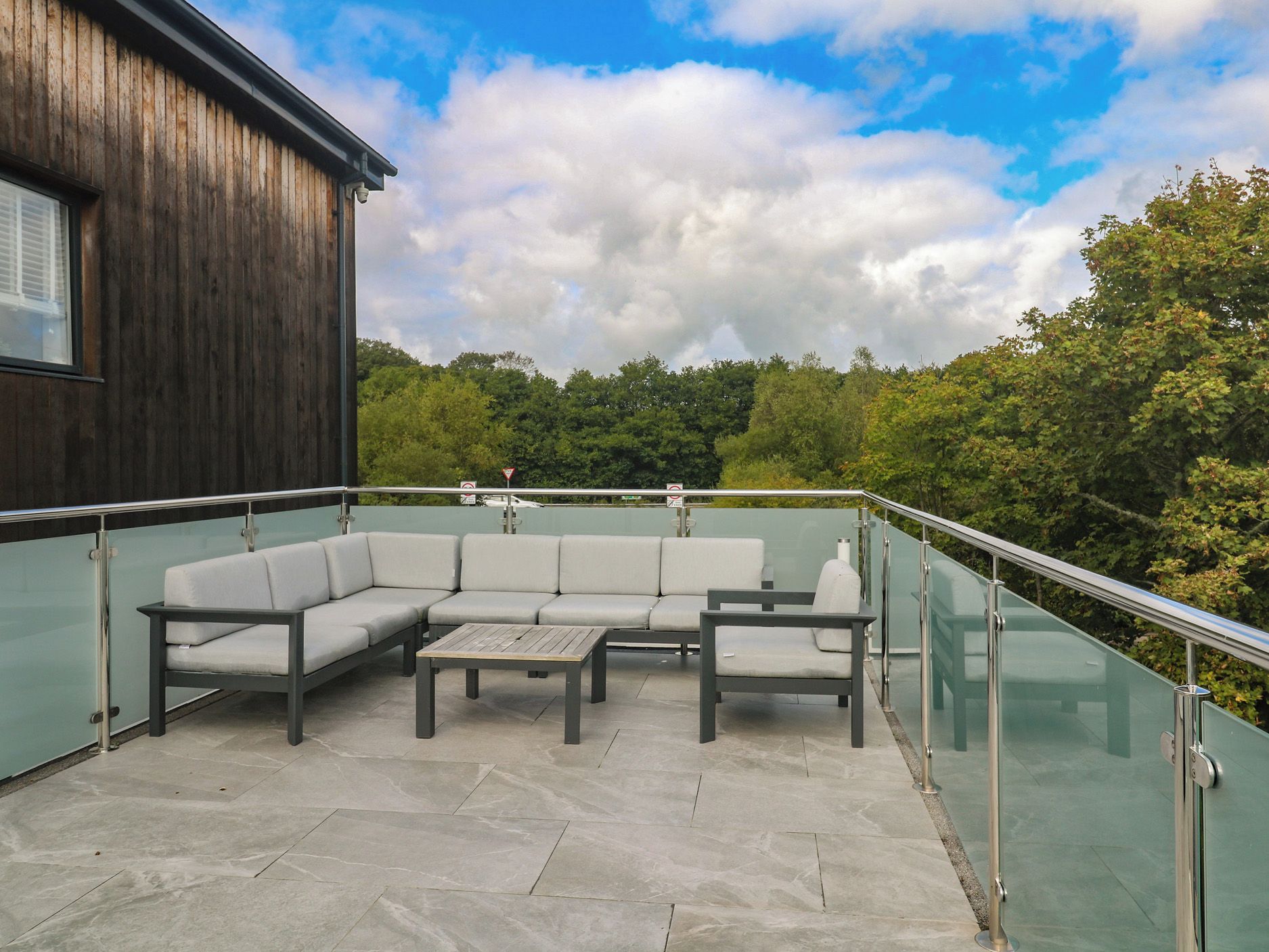 A modern outdoor seating area with a glass balustrade, a wooden building, and a view of a forested landscape.