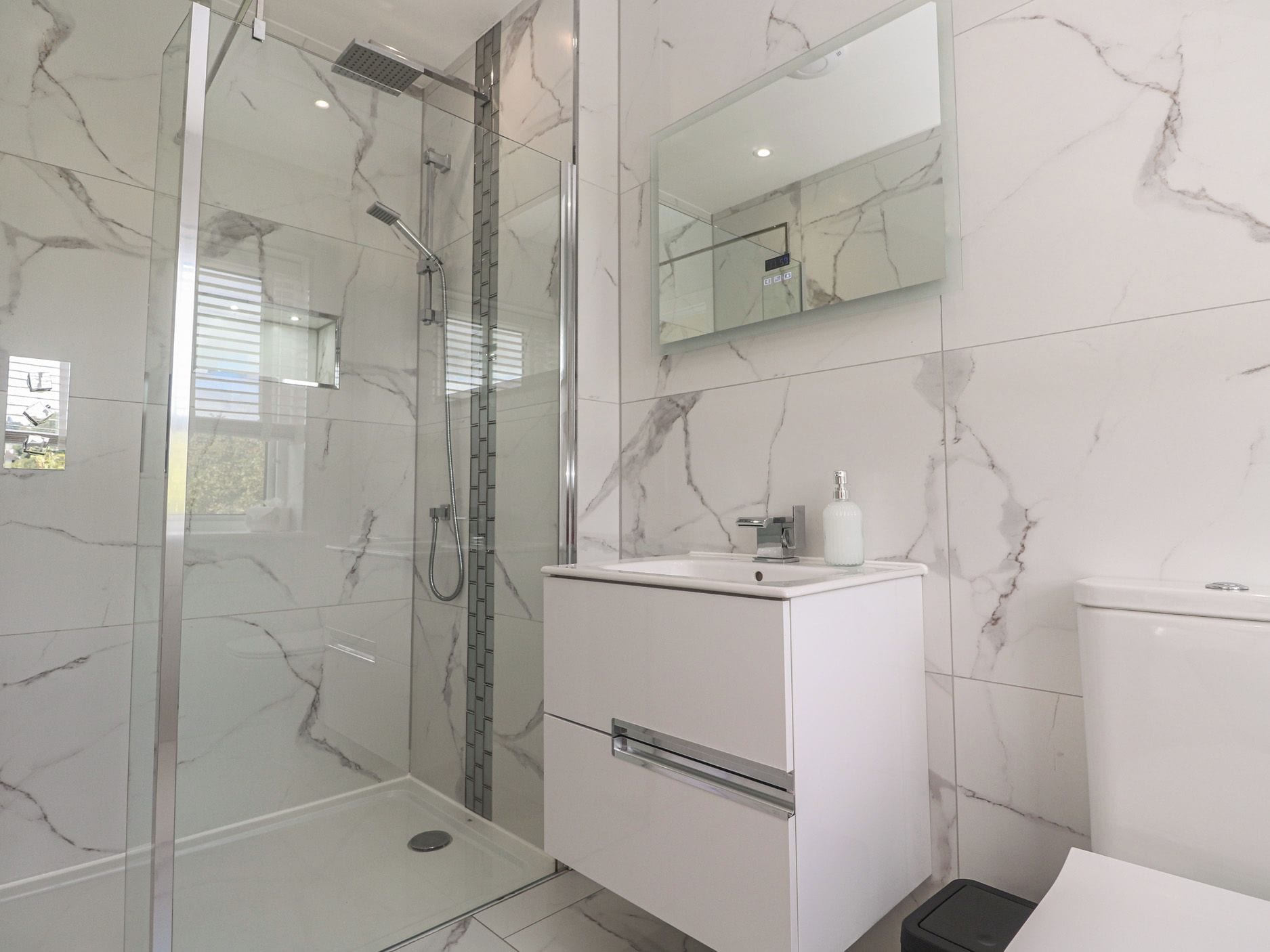 Un baño moderno con ducha de vidrio, tocador blanco con espejo y paredes con diseño de mármol.