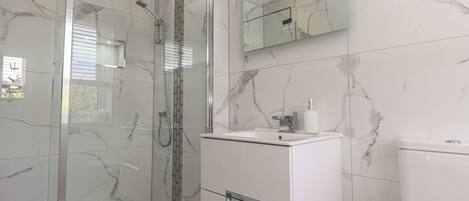 Un baño moderno con ducha de vidrio, tocador blanco con espejo y paredes con diseño de mármol.